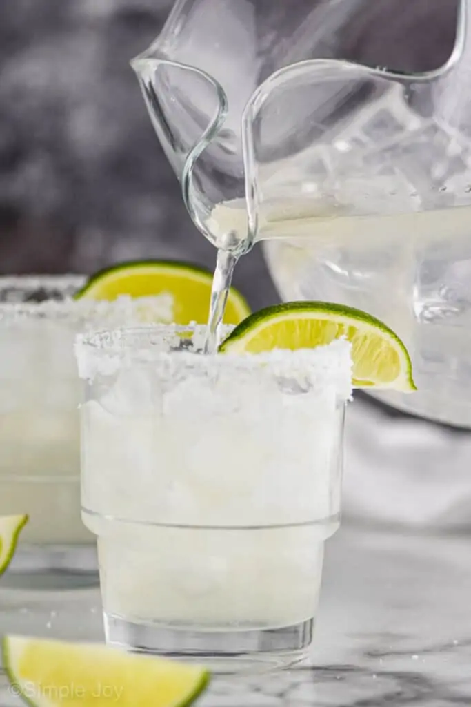 a glass pitcher pouring the best margarita recipe into a glass filled with ice with a salted rim and a lime wedge on the rim, another margarita blurred in the background