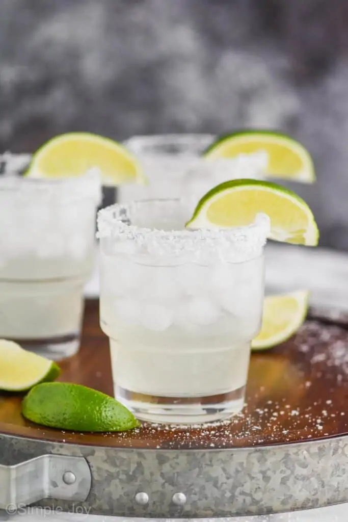 three small tumblers filled with a margarita recipe with salted rims and lime wedges on a wooden tray that has a galvanized metal rim with three lime wedges and lots of spilled kosher salt on the tray