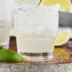 up close straight on view of a tumbler full of a margarita on ice with a salted rim and a lime wedge, two blurred margaritas int he background on a galvanized metal and wooden tray