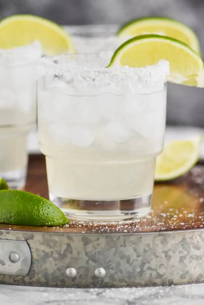 up close straight on view of a tumbler full of a margarita on ice with a salted rim and a lime wedge, two blurred margaritas int he background on a galvanized metal and wooden tray