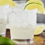 up close straight on view of a tumbler full of a margarita on ice with a salted rim and a lime wedge, two blurred margaritas int he background on a galvanized metal and wooden tray
