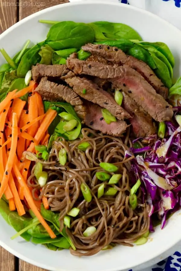 An overhead photo of a bowl of the Soba Noodle Steak Salad that has spinach, carrots, sliced steaks, red cabbage, soba noddles, and topped with black sesame seeds and scallions. 