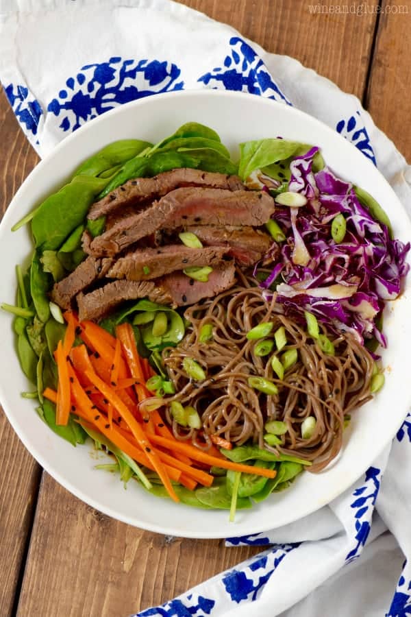 An overhead photo of a bowl of the Soba Noodle Steak Salad that has spinach, carrots, sliced steaks, red cabbage, soba noddles, and topped with black sesame seeds and scallions. 