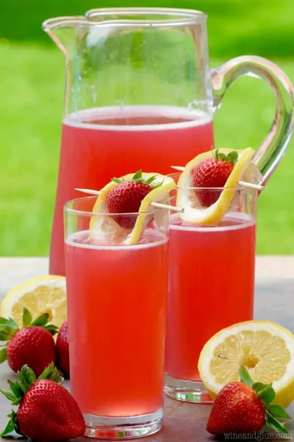 Two glasses of the Strawberry Lemonade Beergaritas are in front of a pitcher of the drink which has a pink red tint. 
