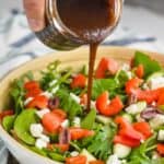 a hand pouring balsamic salad dressing onto a salad with mixed greens, tomatoes, red peppers, feta cheese, and kalamata olives