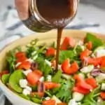 a hand pouring balsamic salad dressing onto a salad with mixed greens, tomatoes, red peppers, feta cheese, and kalamata olives