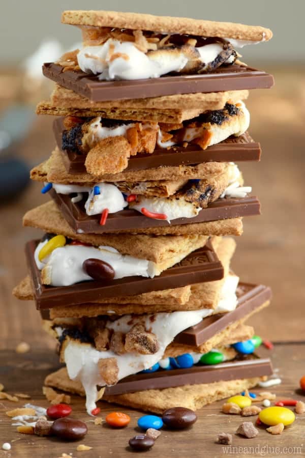 The Candy Coated S'mores is stacked on top of each other showing the cut chocolate bar, M&Ms, sprinkles, Butterfinger, melted marshmallow, and graham crackers. 