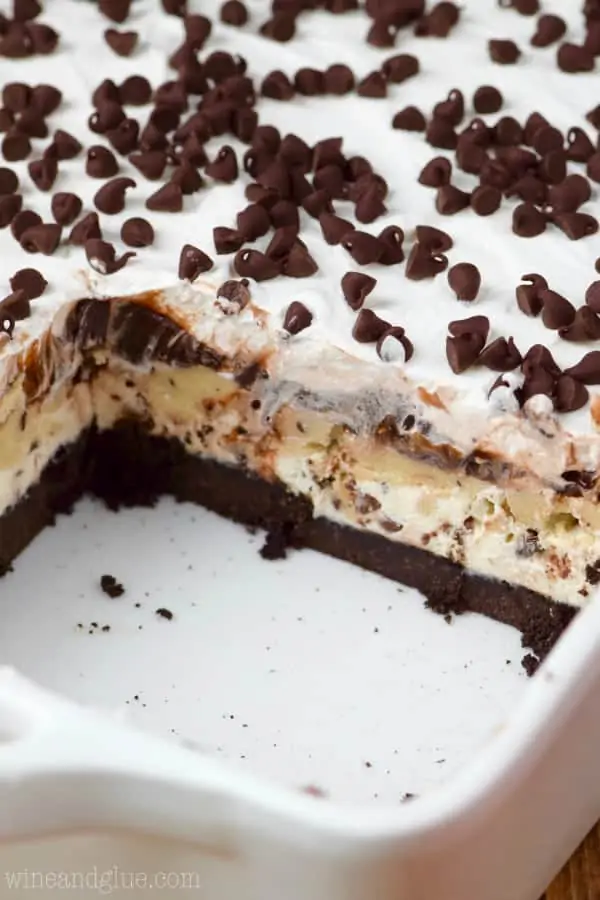 In a ceramic casserole dish, the Cookie Dough Lush has a little rectangular slice cut our showing the distinct layers of the Oreo crust, cookie dough filling, and a white fluffy frosting with mini chocolate chips. 