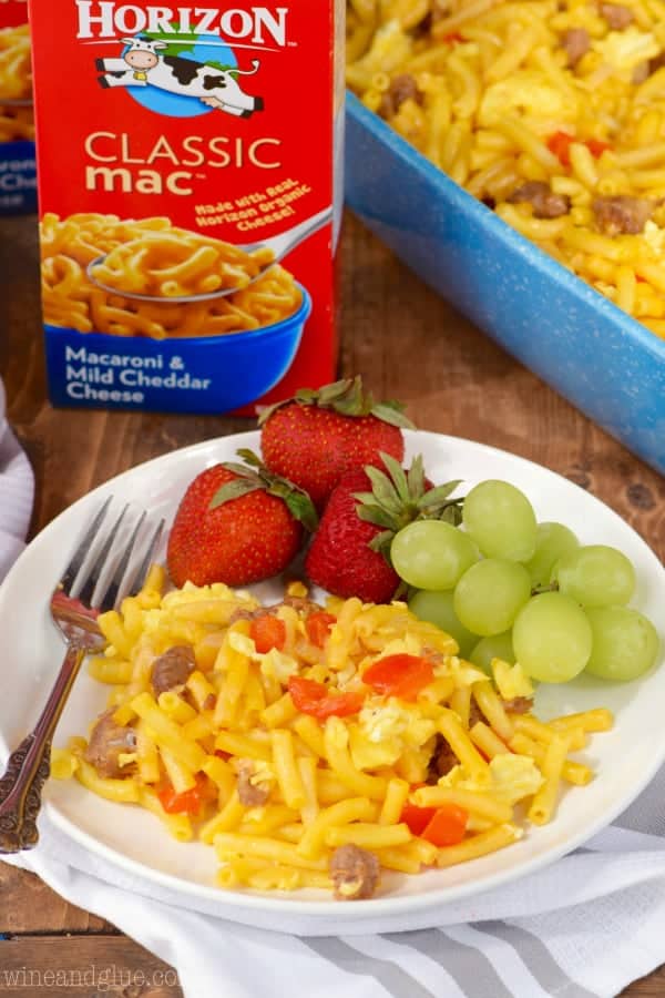On a white plate, the Mac N' Cheese Breakfast Casserole has chunks of sausage, eggs, and sliced red peppers with a side of strawberries and grapes. 
