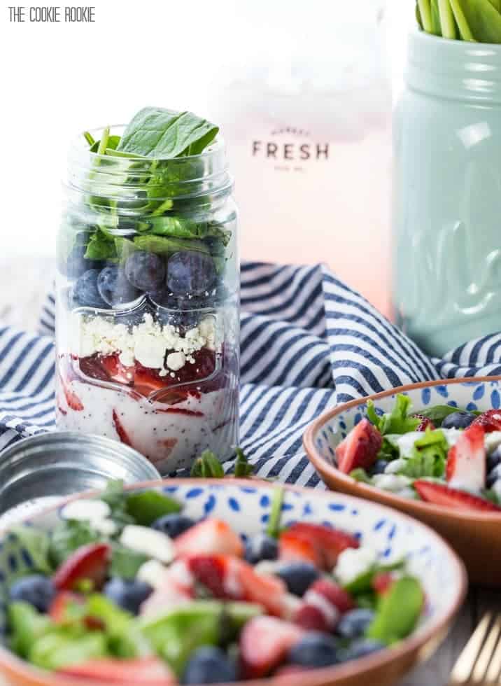 The Red, White, and Blue Mason Jar Salad has distinct layers: a white sauce bottom, cut strawberries, cheese, blueberries, and spinach. 