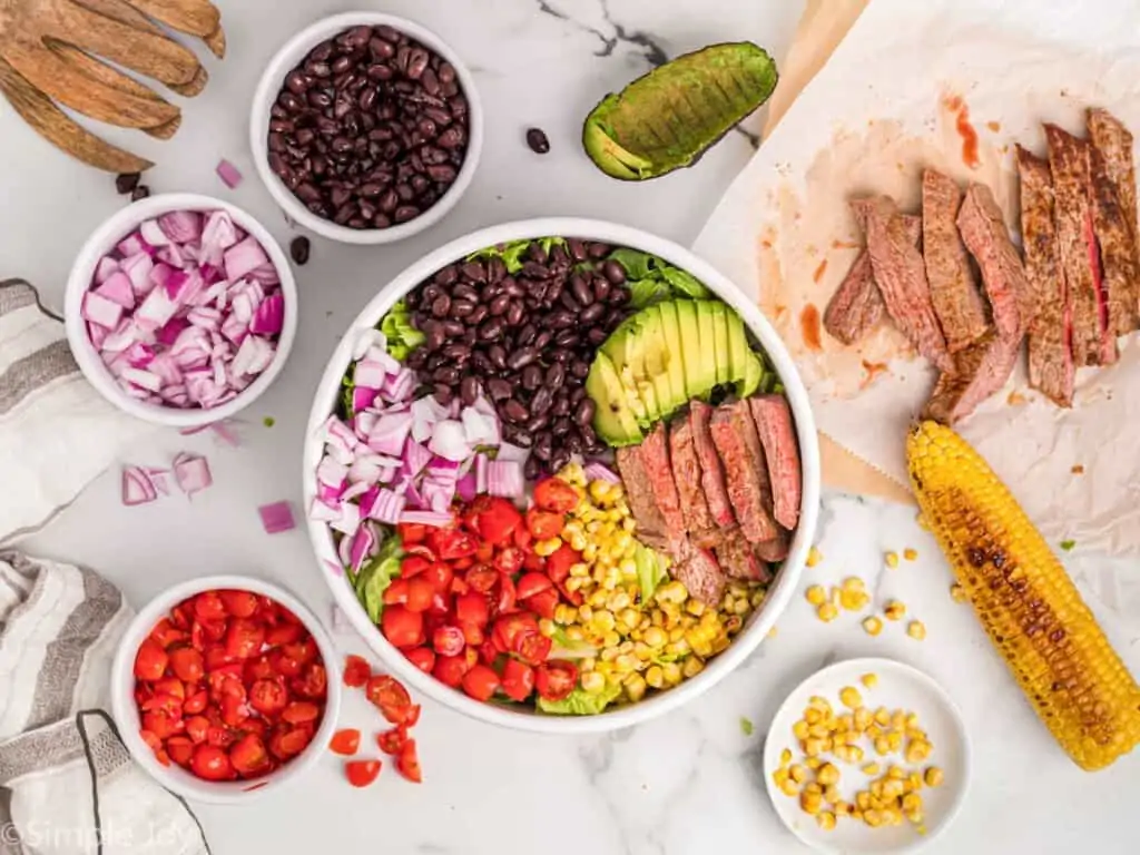 overhead photo of ingredients for a steak salad in a bowl with more ingredients around the bowl