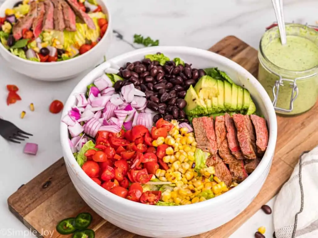 side view of a salad with steak in a big white bowl with dressing next to it