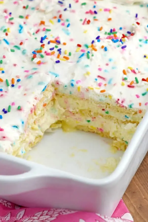 In a white dish, the No Bake Birthday Cake Lasagna has a pudding filling with a Belgium crips in between and topped with frosting and rainbow sprinkles.  