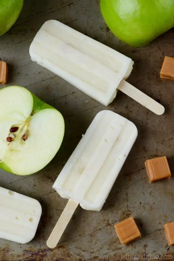 On a baking sheet, the Lighter Caramel Apple Pipe Pops are surrounded by Green apples and caramel cubes. 