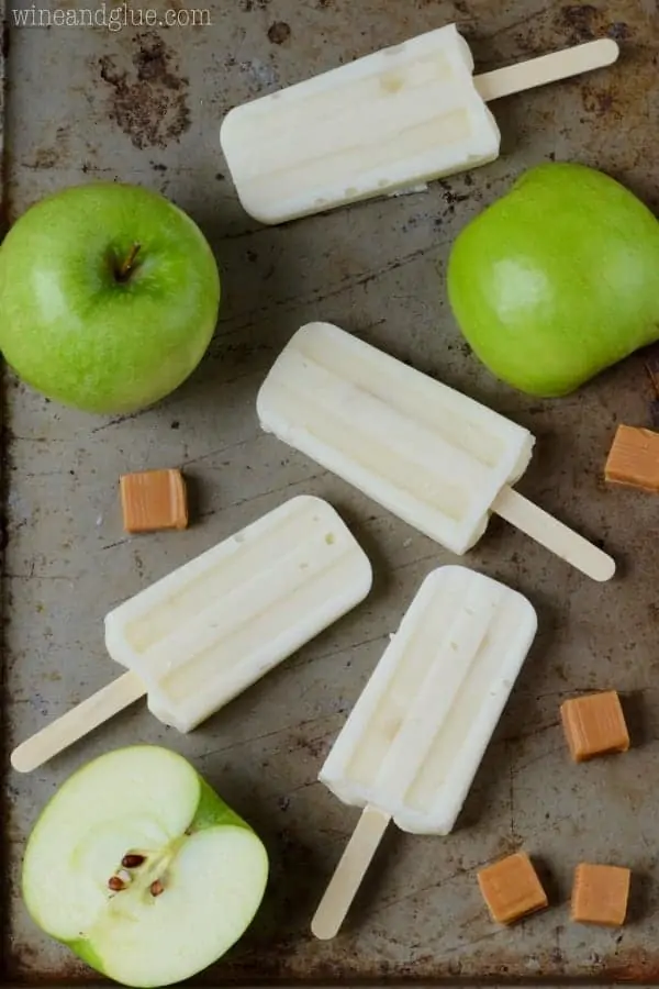 On a baking sheet, the Lighter Caramel Apple Pipe Pops are surrounded by Green apples and caramel cubes. 