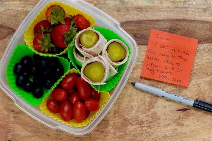 A lunch box filled with strawberries, Salami Pickles Roll, cherry tomatoes, and olives next to a lunch note. 