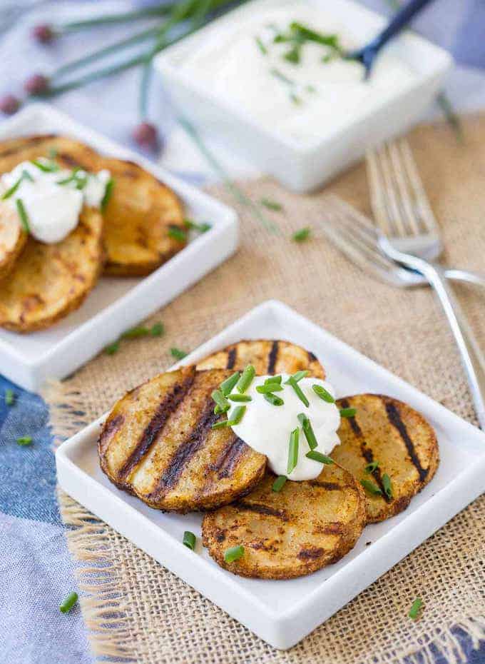 On a small white dish, cut into circular slices the Seasoned Grilled Potatoes is topped with a dollop of sour cream and chives. 