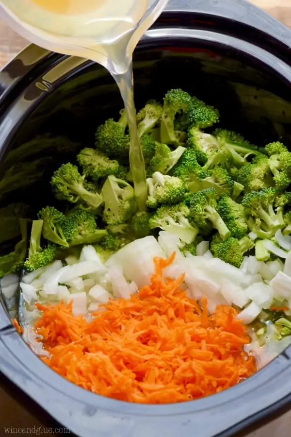 chicken stock being poured into a slow cooker with broccoli, onion, and carrots