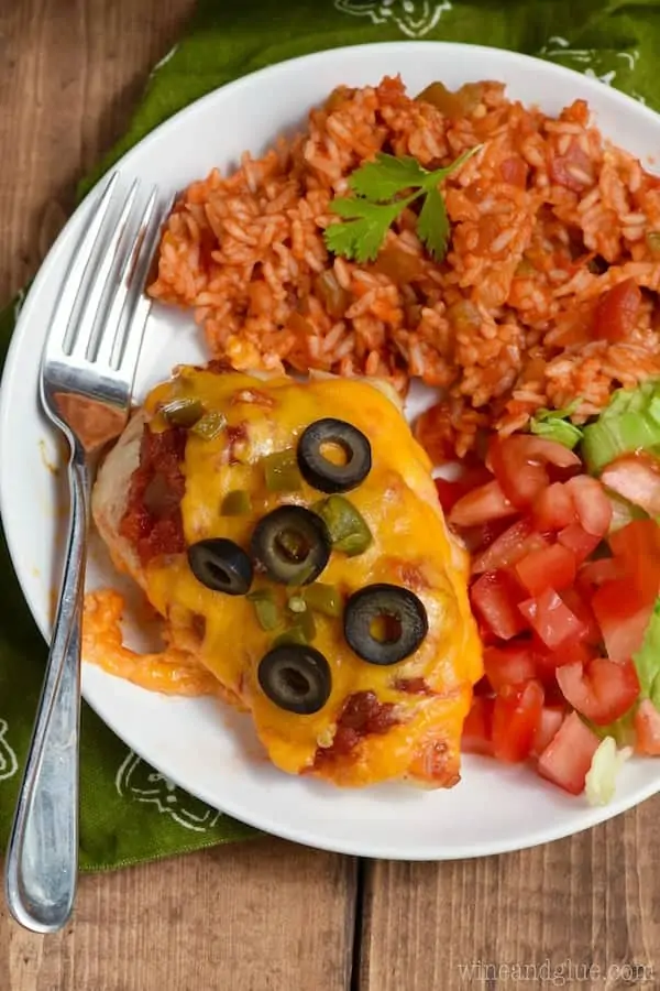 On a white plate, the Stuffed Taco Chicken is covered with melted cheese, olives, and minced jalapenos with sides of cut tomatoes, lettuce, and rice. 