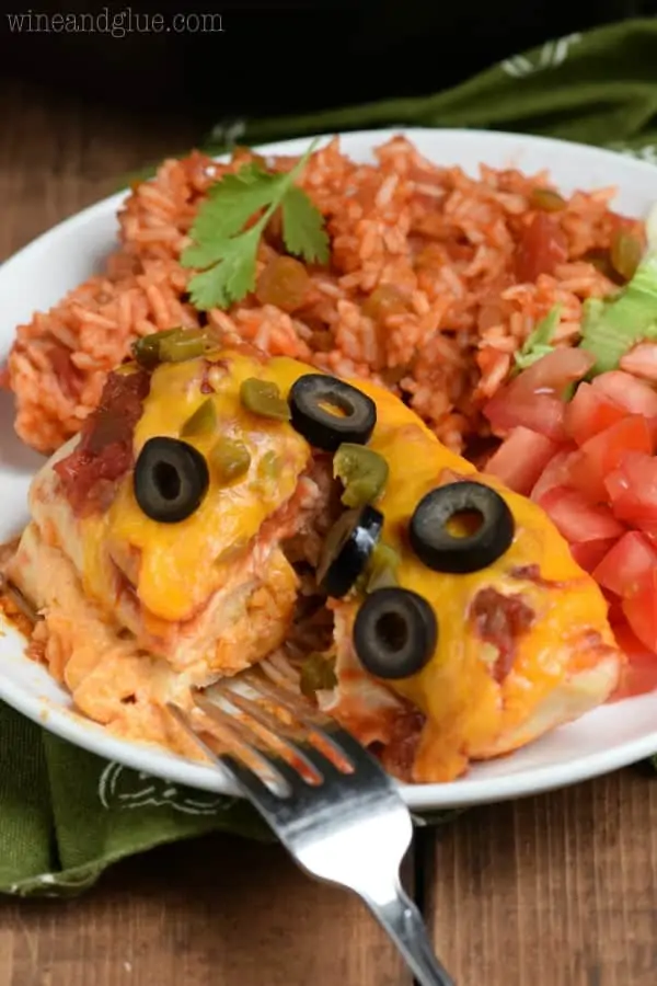 On a white plate, the Stuffed Taco Chicken is covered with melted cheese, olives, and minced jalapenos with sides of cut tomatoes, lettuce, and rice. 