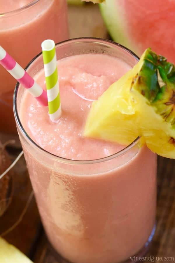 In a tall glass, the Watermelon Pina Colada has a pink hue with a sliced triangular pineapple on the rim. 
