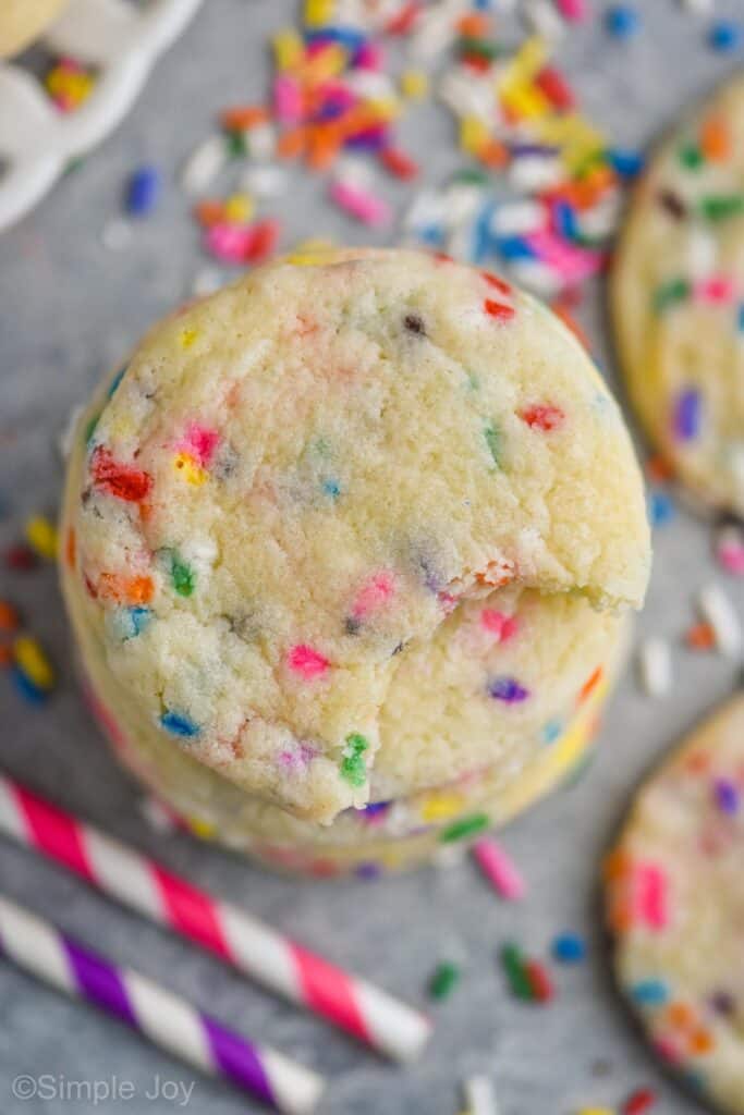 overhead picture of a stack of sugar cookies that has sprinkles added to them, with more sugar cookies, sprinkles, and striped straws blurred beneath