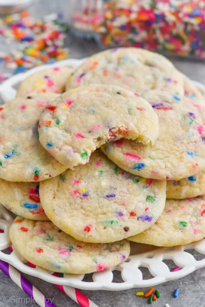 close up of a plate of chewy sugar cookies that have sprinkles added to them, with a bite missing out of the top cookie and a tipped over bottle of sprinkles in the background