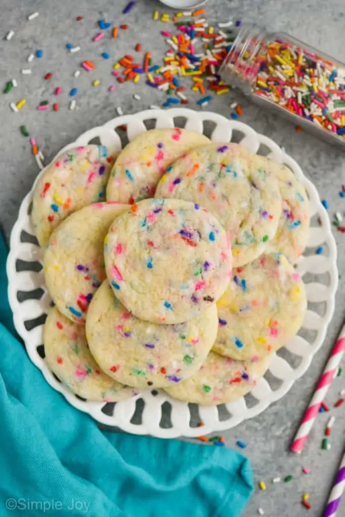 overhead view of a white plate with funfetti cookie recipe, a cloth teal napkin near the plate, and a tipped over glass bottle of sprinkles on it's side