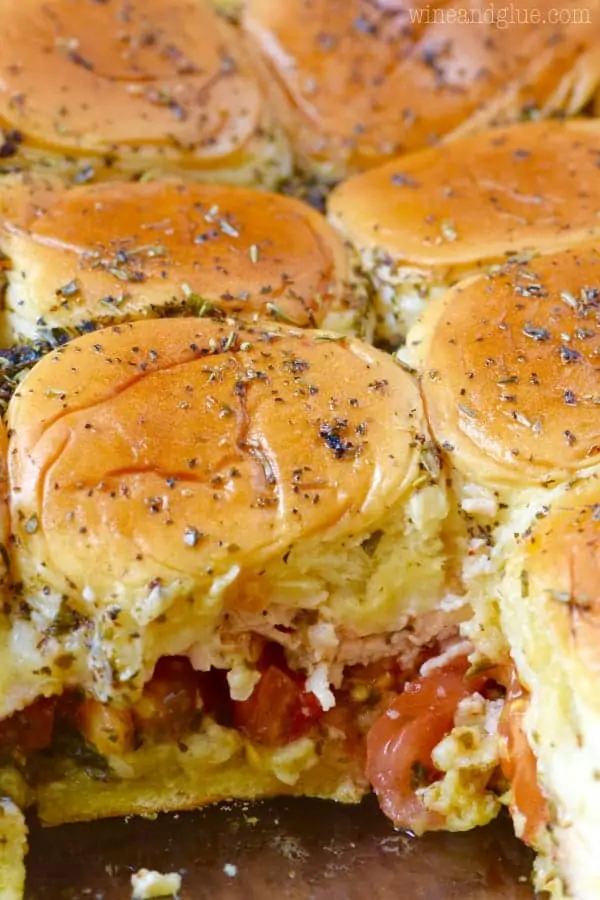 A closeup photo of the Italian Peso Sliders in the casserole dish and one is taken out showing the distinct layers of pesto, tomatoes, turkey, and cheese. 
