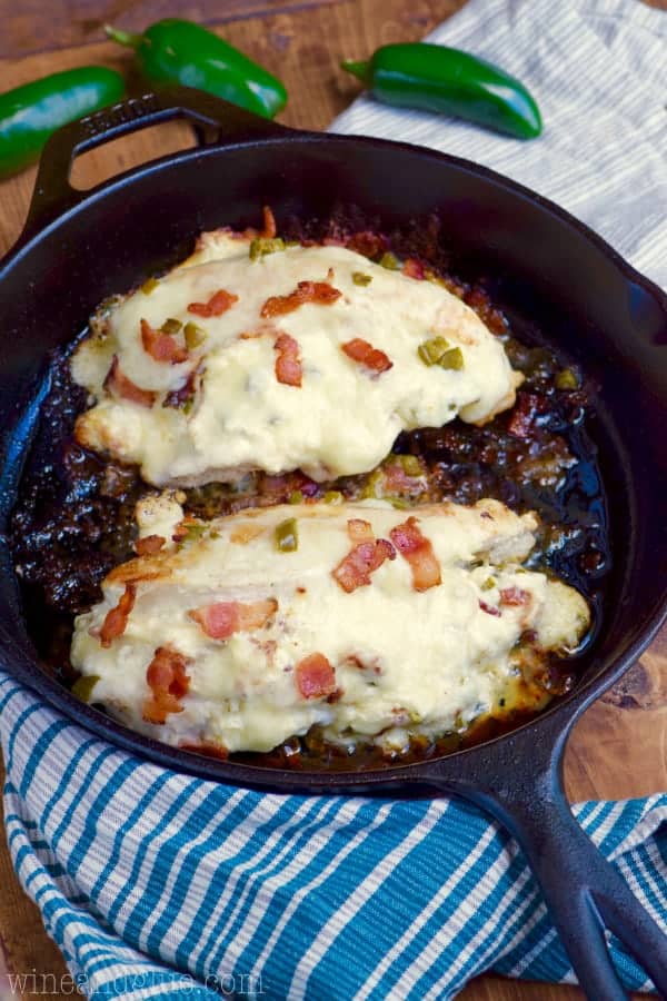 A cast iron skillet has two Jalapeno Popper Stuffed Chicken is covered with melted cheese and topped with bacon and jalapeno slices. 