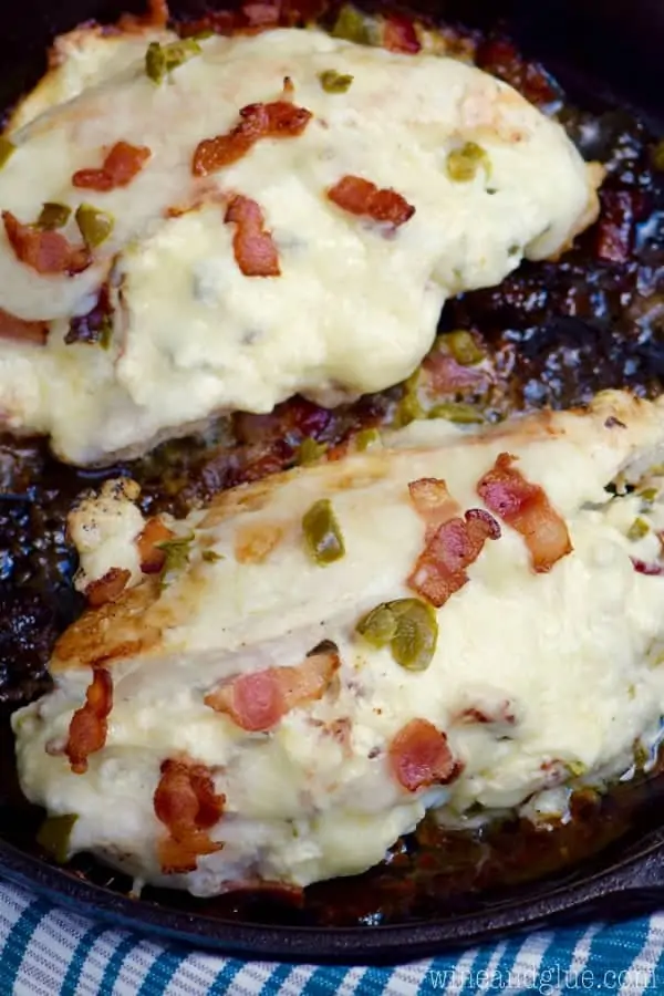 A cast iron skillet has two Jalapeno Popper Stuffed Chicken is covered with melted cheese and topped with bacon and jalapeno slices. 