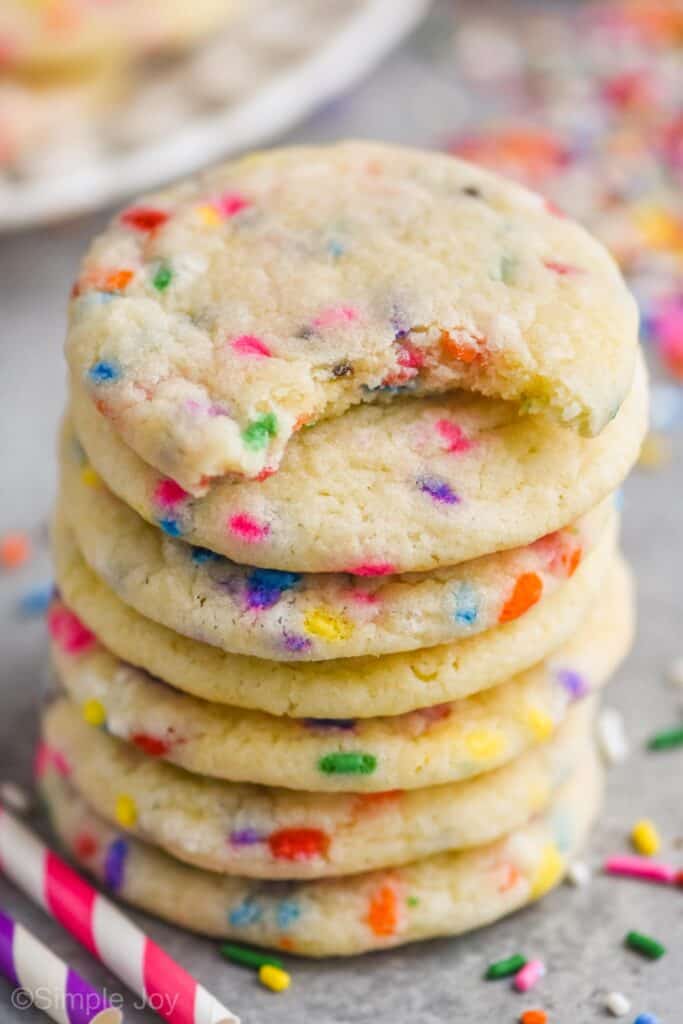 stack of sugar cookies with sprinkles with the top one missing a bite, sprinkles around the stack, and two striped straws