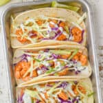overhead view of three shrimp tacos in corn tortillas garnished with cilantro cream and green and red cabbage on a small baking tray with lime wedges around them