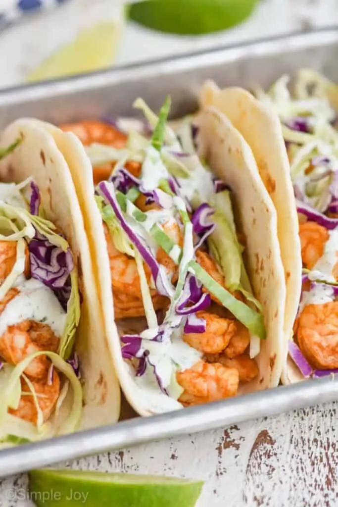 slightly pulled back front view of easy shrimp tacos that are garnished with red and green cabbage on a rimmed baking sheet with two lime wedges in the background and one in the foreground