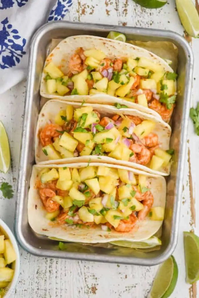 overhead view of three shrimp tacos on a rimed small metal baking sheet, topped with pineapple salsa with lime wedges on the white wood surface around them