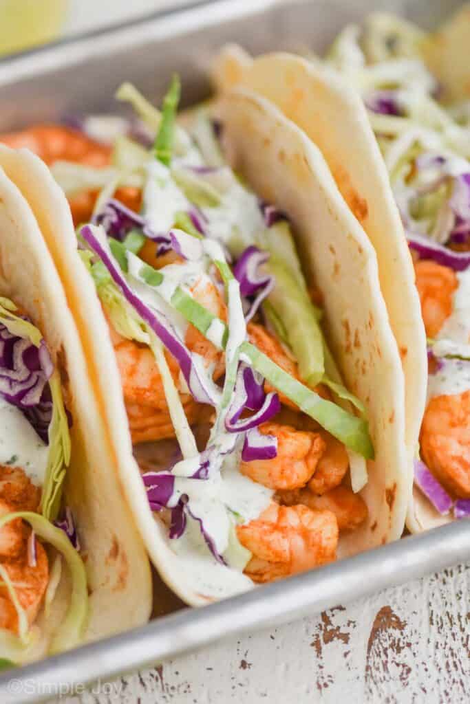 close up front view of a shrimp taco recipe with tacos on either side, tacos are garnished with red and green cabbage and visibly seasoned, they also have cilantro creamy on them and are on a rimed metal baking sheet