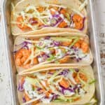 overhead view of three shrimp tacos in corn tortillas garnished with cilantro cream and green and red cabbage on a small baking tray with lime wedges around them - 13