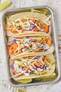 overhead view of three shrimp tacos in corn tortillas garnished with cilantro cream and green and red cabbage on a small baking tray with lime wedges around them