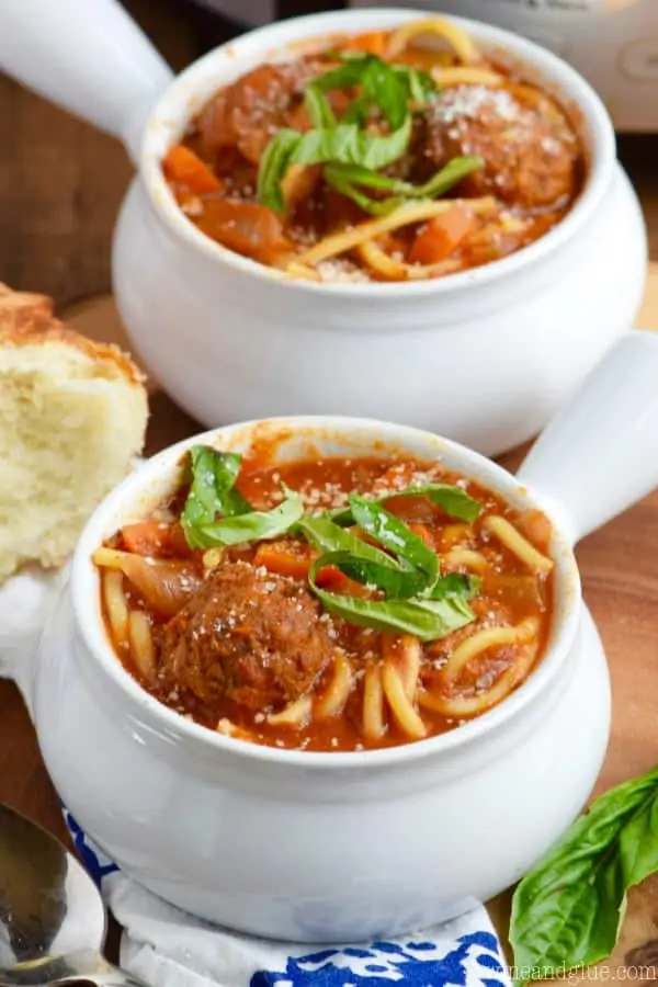In a small white bowl, the Slow Cooker Spaghetti and Meatball Soup is topped with some cheese and basil. 
