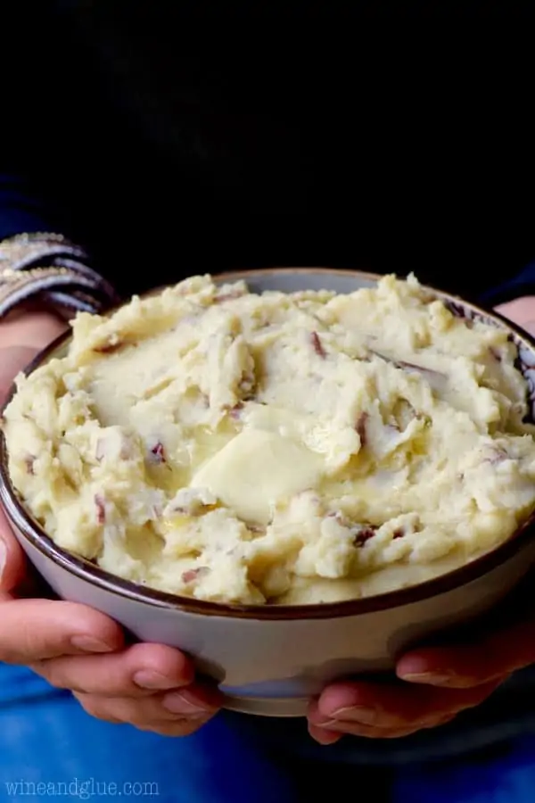 A woman is holding a purple bowl filled with the Cheesy Crock Pot Mashed Potato topped with a single slice of butter. 