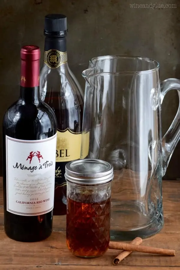 The ingredients of the Cinnamon Sangria is next to a pitcher. (Red wine, honey, cinnamon sticks, and brandy)