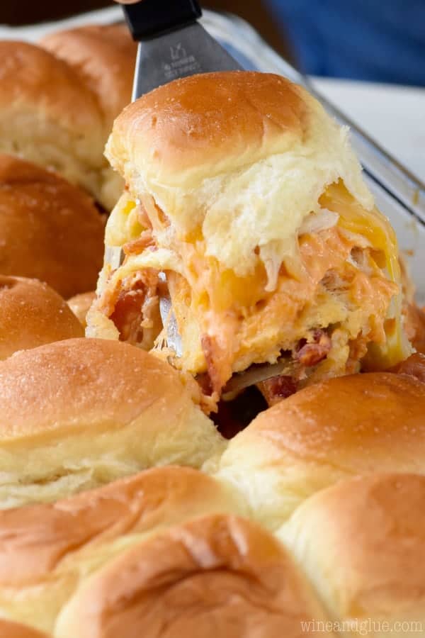 In a casserole dish, a single Sriracha Bacon Slider is being taken out by a spatula showing the melted cheese oozing out. 
