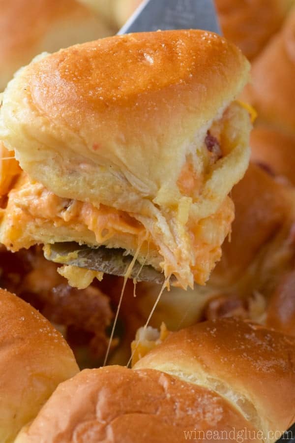 In a casserole dish, a single Sriracha Bacon Slider is being taken out by a spatula showing the melted cheese oozing out. 