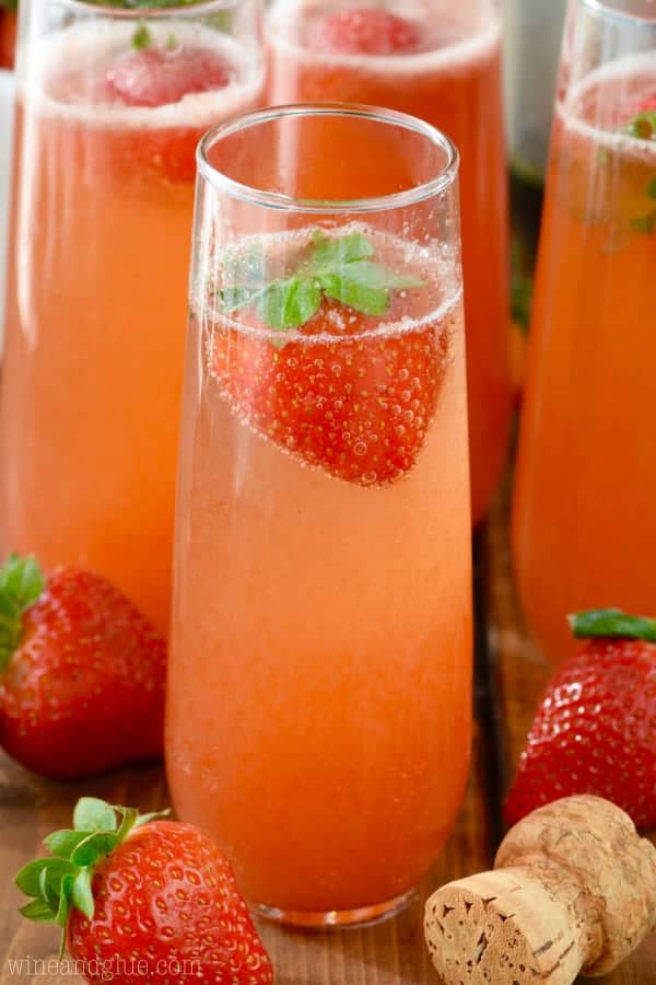 In a champagne glass, the Strawberry Bellinis has a gradient red color, bubbly, and a single strawberry in the glass. 