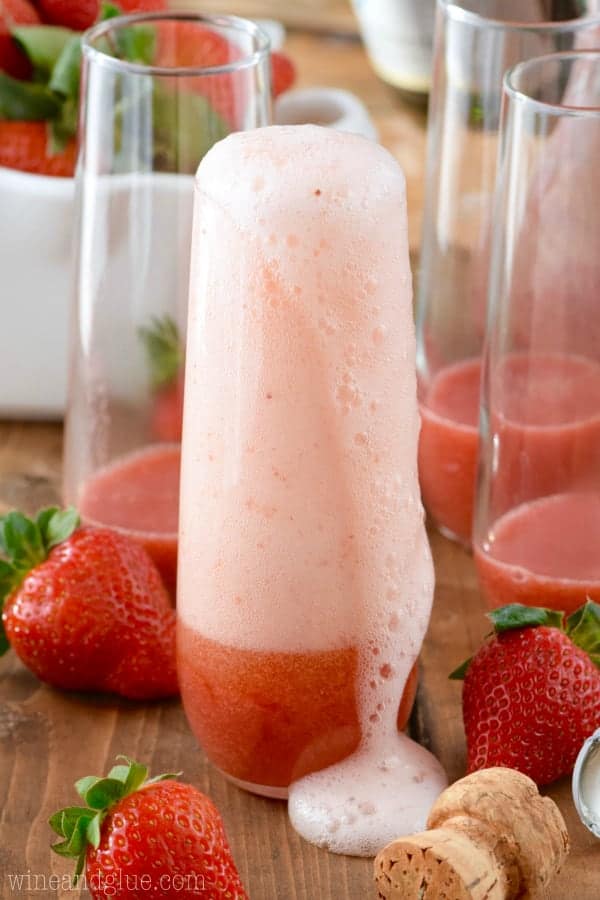 Champagne is fizzing out of a tall glass of Strawberry Bellinis with strawberry puree. 