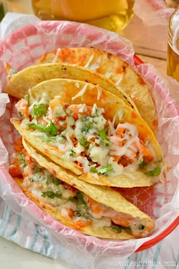 In a lunch basket, three Buffalo Baked Tacos has a bright red color from the Buffalo sauce and melted cheese and topped with chives. 