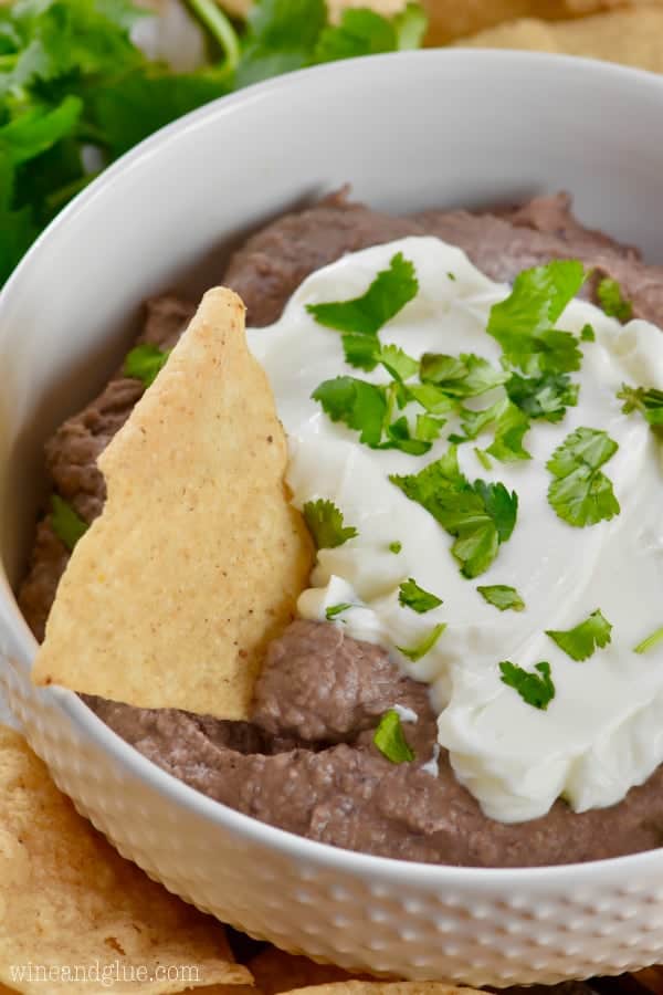 a tortilla chip being dipped into the black bean dip in a white bowl with sour cream and cilantro