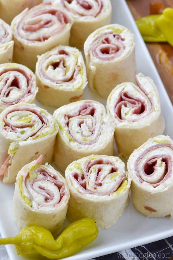On a white plate, the Italian Pinwheels are in rows. The interior of the Italian Pinwheels show the fillings of pepperoncini, cream cheese, and some deli meat. 