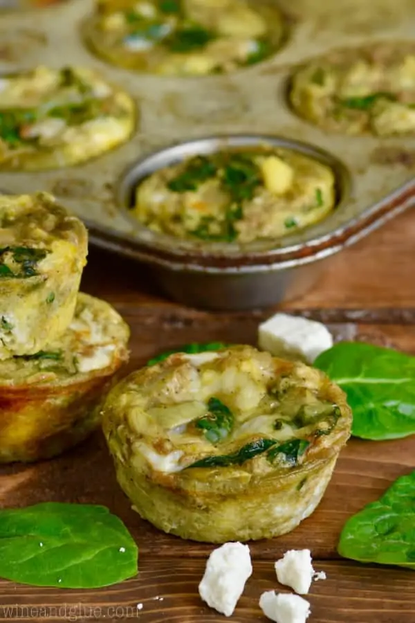 A single Spinach and Artichoke Egg Muffin in front of the muffin tin and surrounded by feta cheese and spinach.