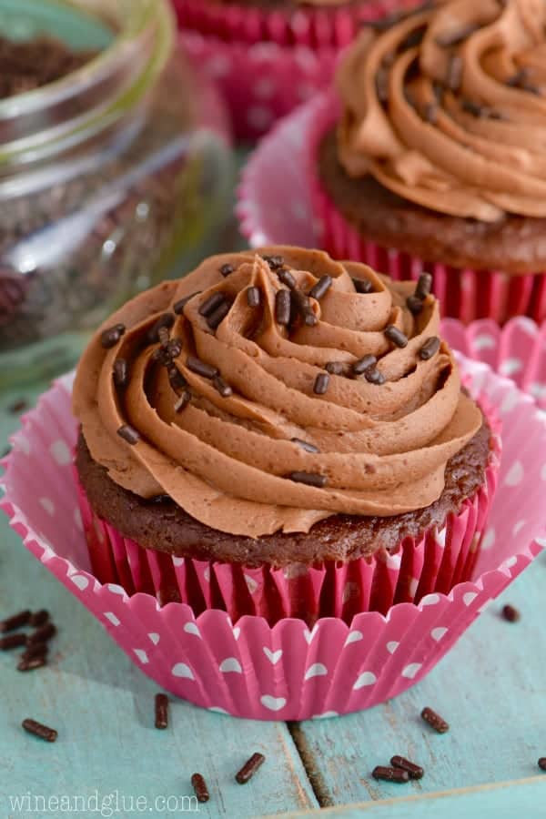 A chocolate muffin in a Valentine's day wrapper has a Chocolate Buttercream Frosting with chocolate sprinkles. 